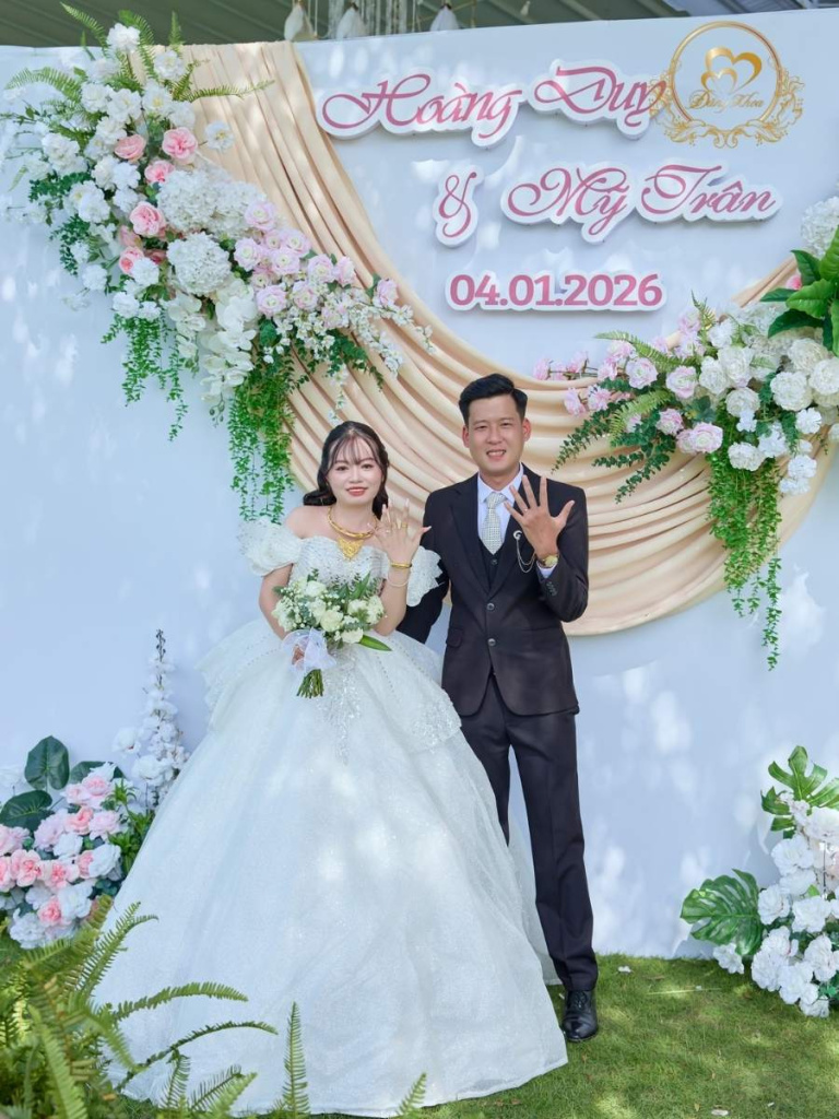 Cô dâu chú rể khoe nhẫn cưới hạnh phúc bên backdrop hoa tone hồng 