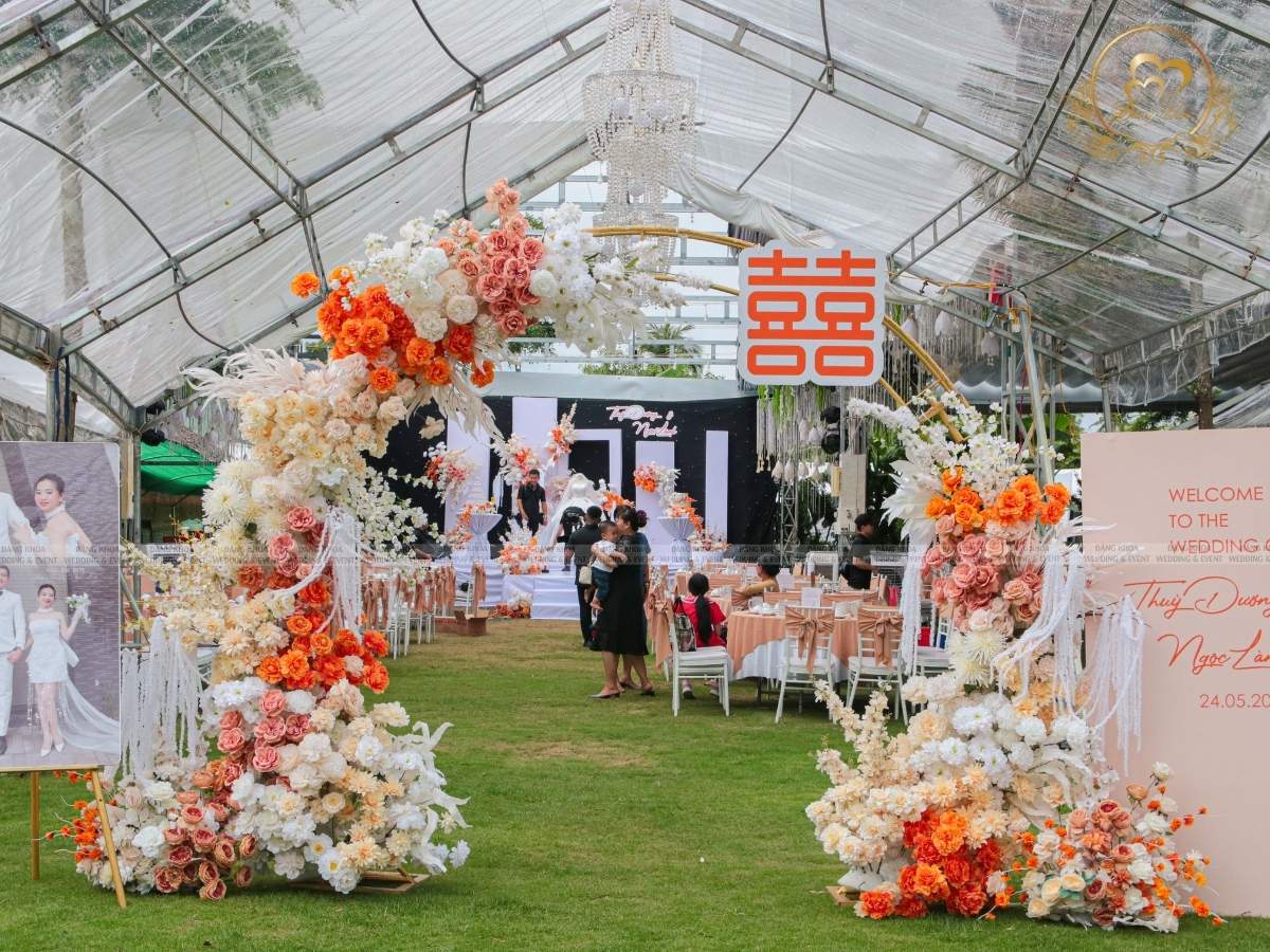 Lối dẫn vào tiệc cưới trang trí cổng hoa và chữ Song Hỷ tone cam nổi bật.