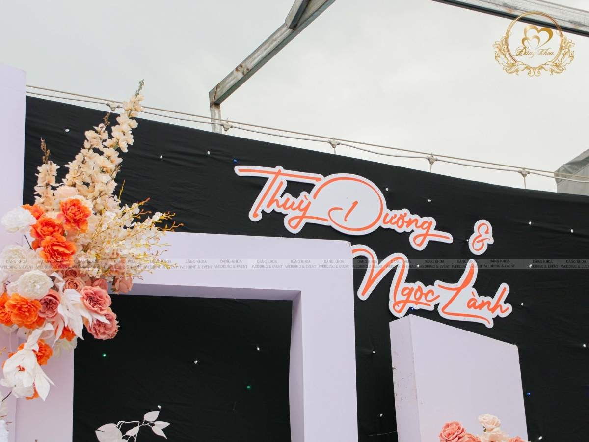 Cận cảnh backdrop sân khấu đám cưới tone trắng cam với tên cố dâu chú rể nổi bật.