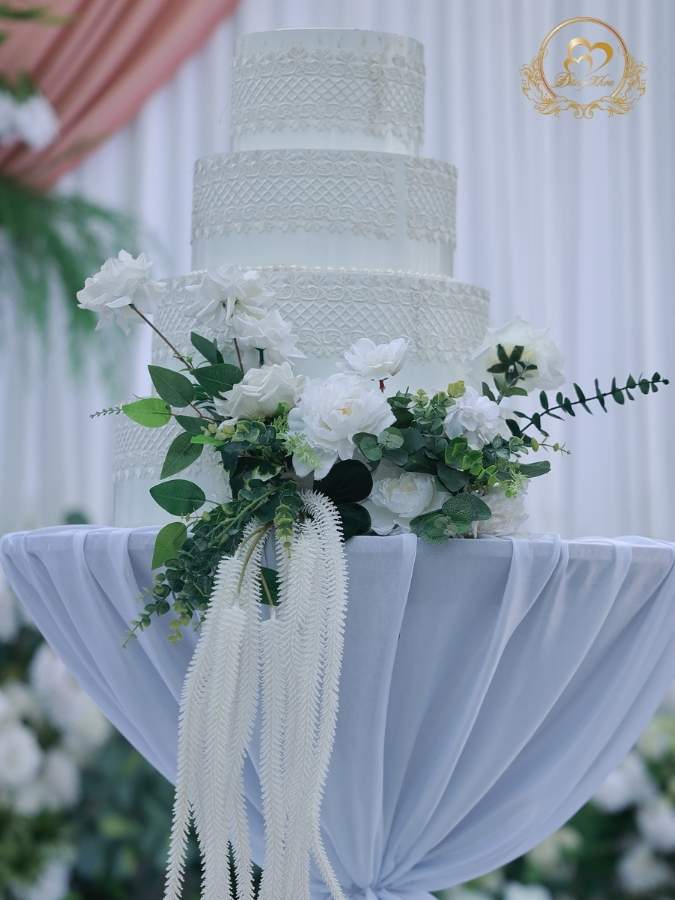 Bánh kem cưới 4 tầng màu trắng trang trí hoa lụa cao cấp và họa tiết ren sang trọng.
